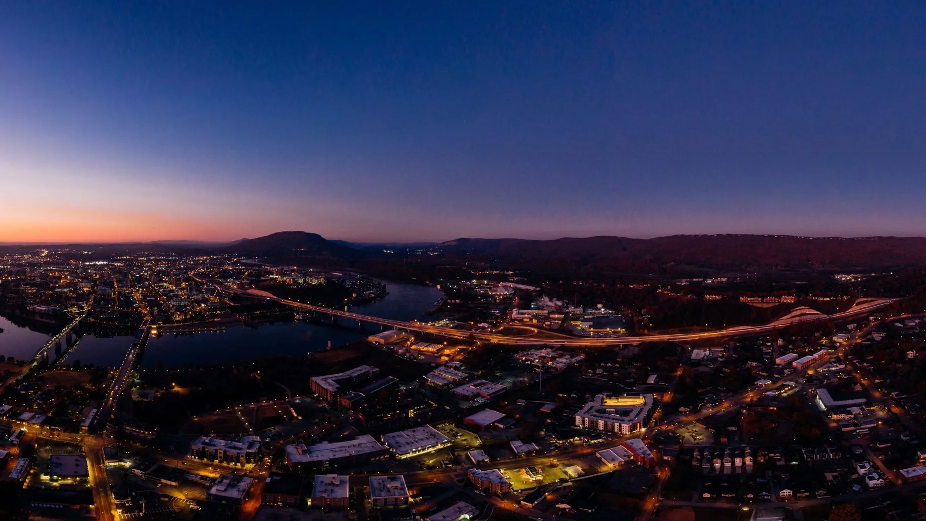 Aerial view of a city at dusk: lit streets and bridges over a river, with hills against an orange horizon